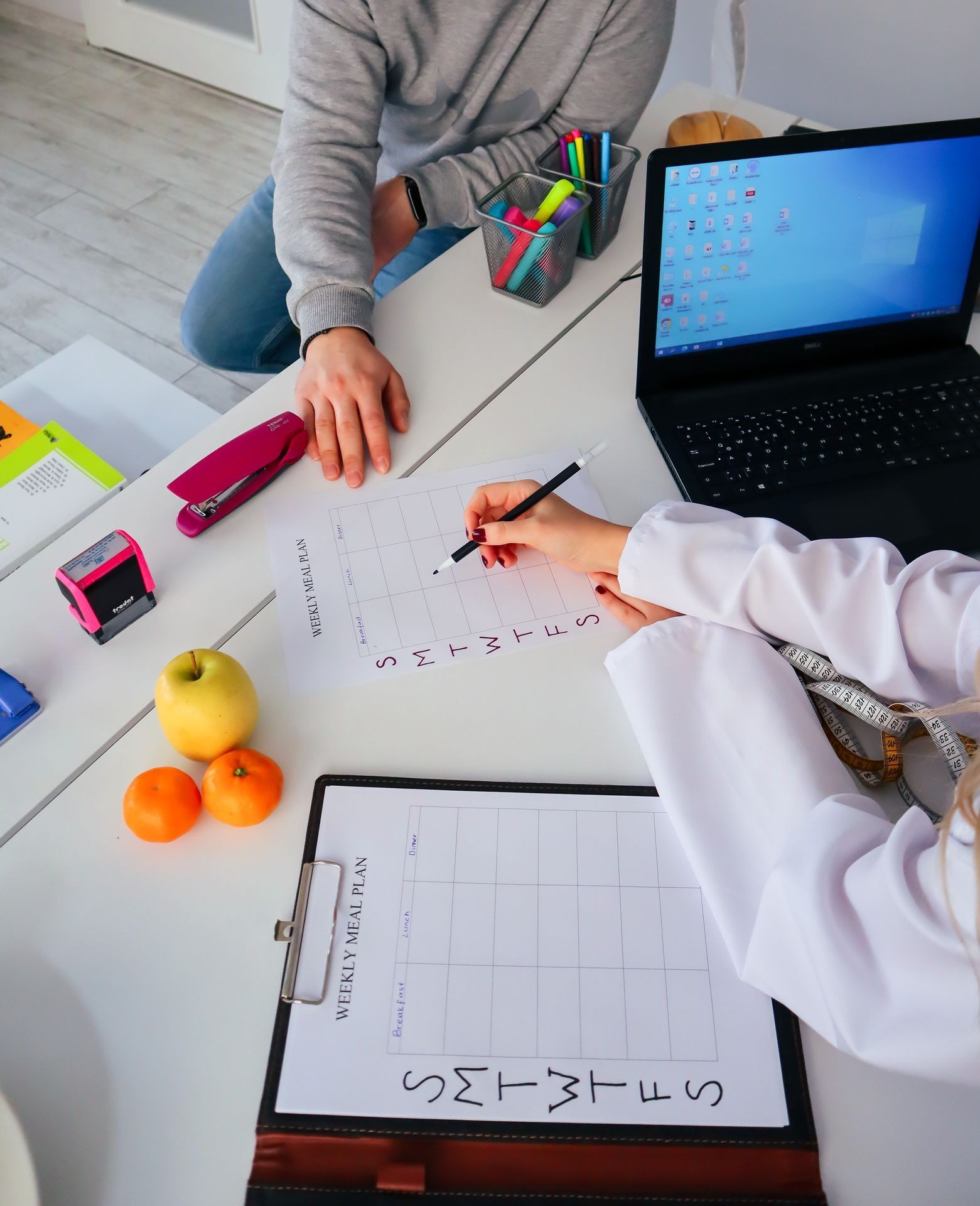 Person planning a weekly meal schedule at a desk with a laptop, fruits, and office supplies.