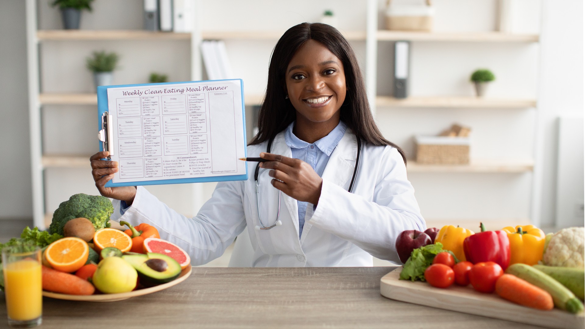 Right nutrition concept. Happy black nutritionist showing weekly diet plan, sitting at desk with fruits and vegetables